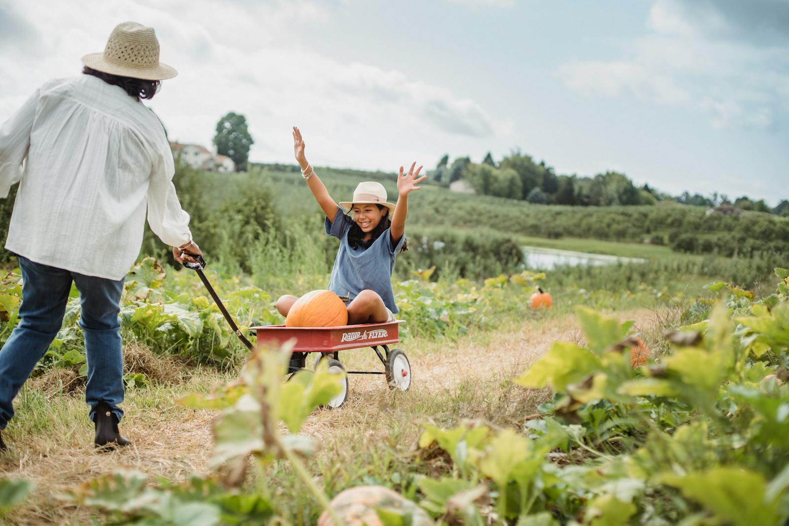 Full body of cheerful young girl in cart with pumpkin enjoying time with mother in countryside